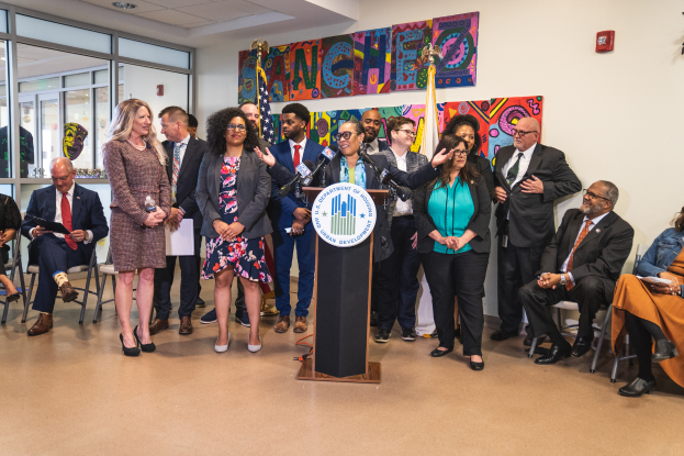 Eine Gruppe von Menschen steht und sitzt in einem Raum mit einem Podium, einem Mikrofon und einer Tafel mit der Aufschrift "Sancho City Councilman Joins City Council", mit Flaggen, einem Feuerlöscher und Deckenleuchten an der Rückwand.