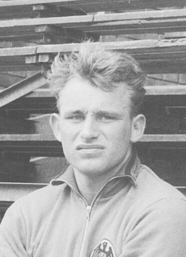 Schwarzes und weißes Foto eines Mannes in einer Jacke, der vor einem Haufen Holzplanken steht, der als der legendäre deutsche Fußballspieler gilt, der im Alter von 89 Jahren verstorben ist.