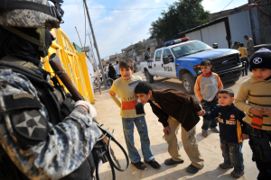 Eine Gruppe von Kindern vor einem Polizisten mit einer Waffe, mit Fahrzeugen, Menschen, Fahrrädern und Bäumen im Hintergrund unter einem bewölktem Himmel.