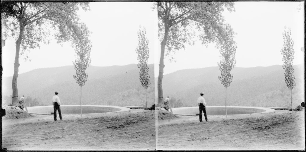 Schwarz-weiß-Foto eines Mannes, der vor einem Teich mit Bäumen, Hügeln und einem klaren Himmel steht, umgeben von üppiger Vegetation.