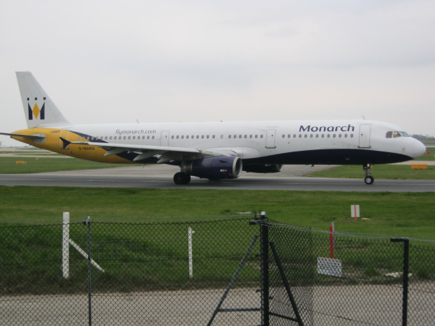 Ein Monarch Airlines Airbus A320-200 auf der Rollbahn, umgeben von Gras, einem Zaun, Bäumen und einem klaren blauen Himmel im Hintergrund.