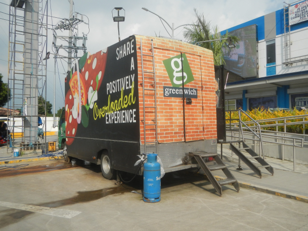 Ein Lkw steht vor einem Gebäude mit einer Seite, auf der ein Schild mit der Aufschrift "Teilen Sie ein positiv überladenes Erlebnis" zu sehen ist. Es gibt Stufen, die zum Lkw führen, einen Zylinder auf dem Boden, Geländer, Strommasten mit Drähten, Laternen, Bäume und einen bewölkten Himmel im Hintergrund.