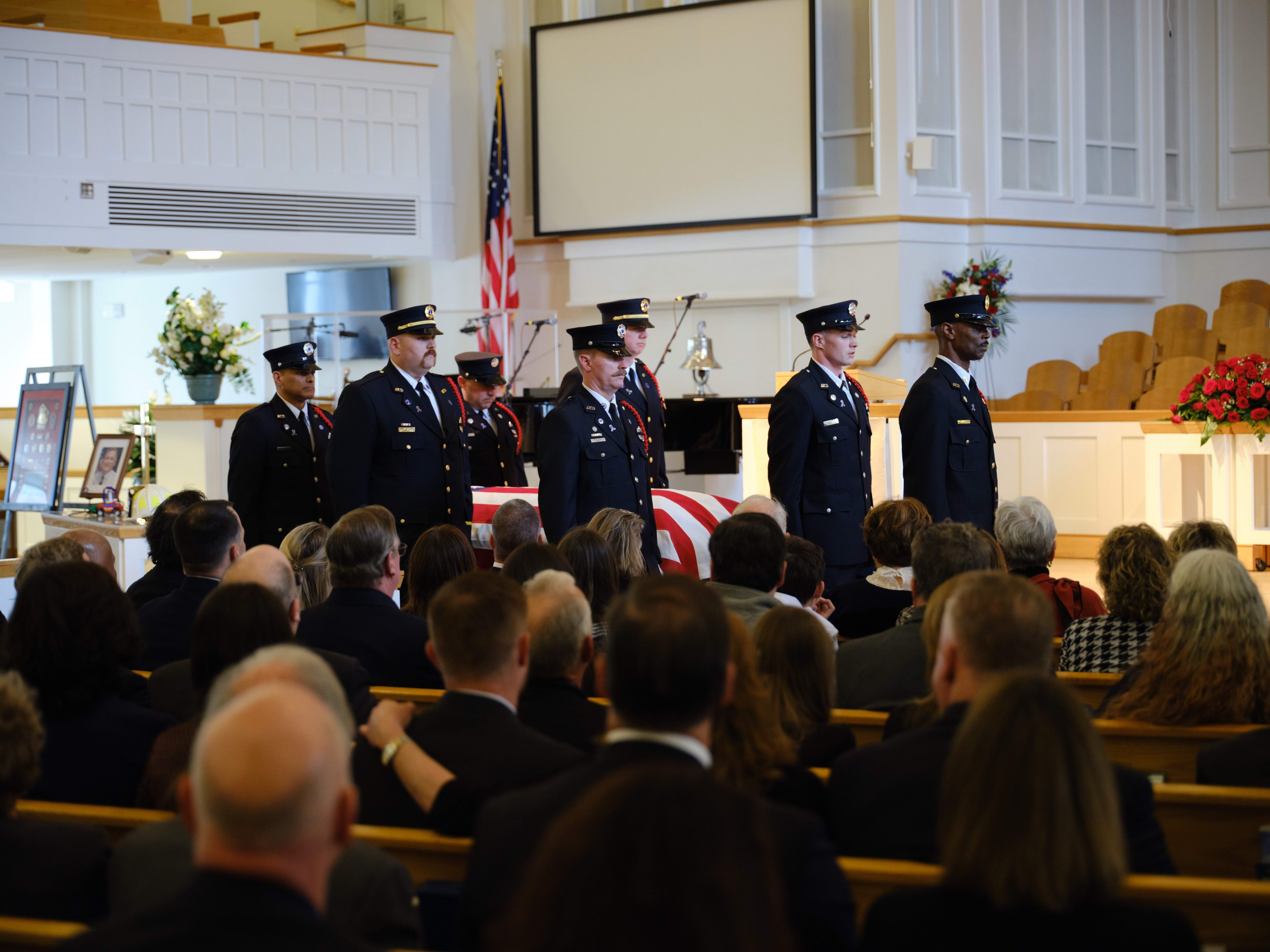 Eine Gruppe uniformierter Personen steht vor sitzenden Trauergästen bei einer Beerdigung, mit Blumensträußen, einer Fahne, einem Bildschirm und einer Wand im Hintergrund.