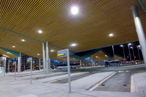 Bus-Haltestelle bei Nacht mit Deckenbeleuchtung, Fahrzeugen auf der Straße, Beschilderung, Menschen und beleuchtete Gebäude im Hintergrund.