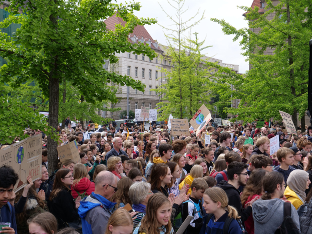 Große Menschenmenge protestiert vor einem Gebäude in Berlin, hölt Schilder, mit Bäumen, Fahrzeugen, einem Lautsprecher und Himmel.