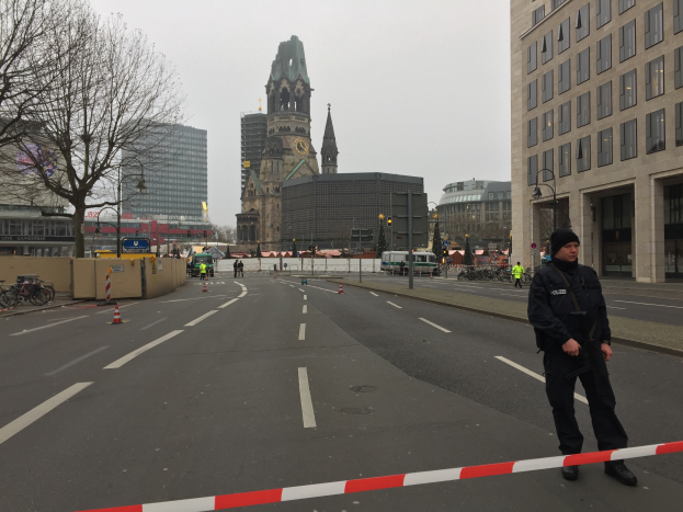Ein Polizeibeamter in schwarzer Uniform und Mütze steht in einer Berliner Straße umgeben von Absperrbaken, Fahrrädern, Laternen, Bäumen und Gebäuden, mit einem Uhrenturm und einem klaren blauen Himmel im Hintergrund.