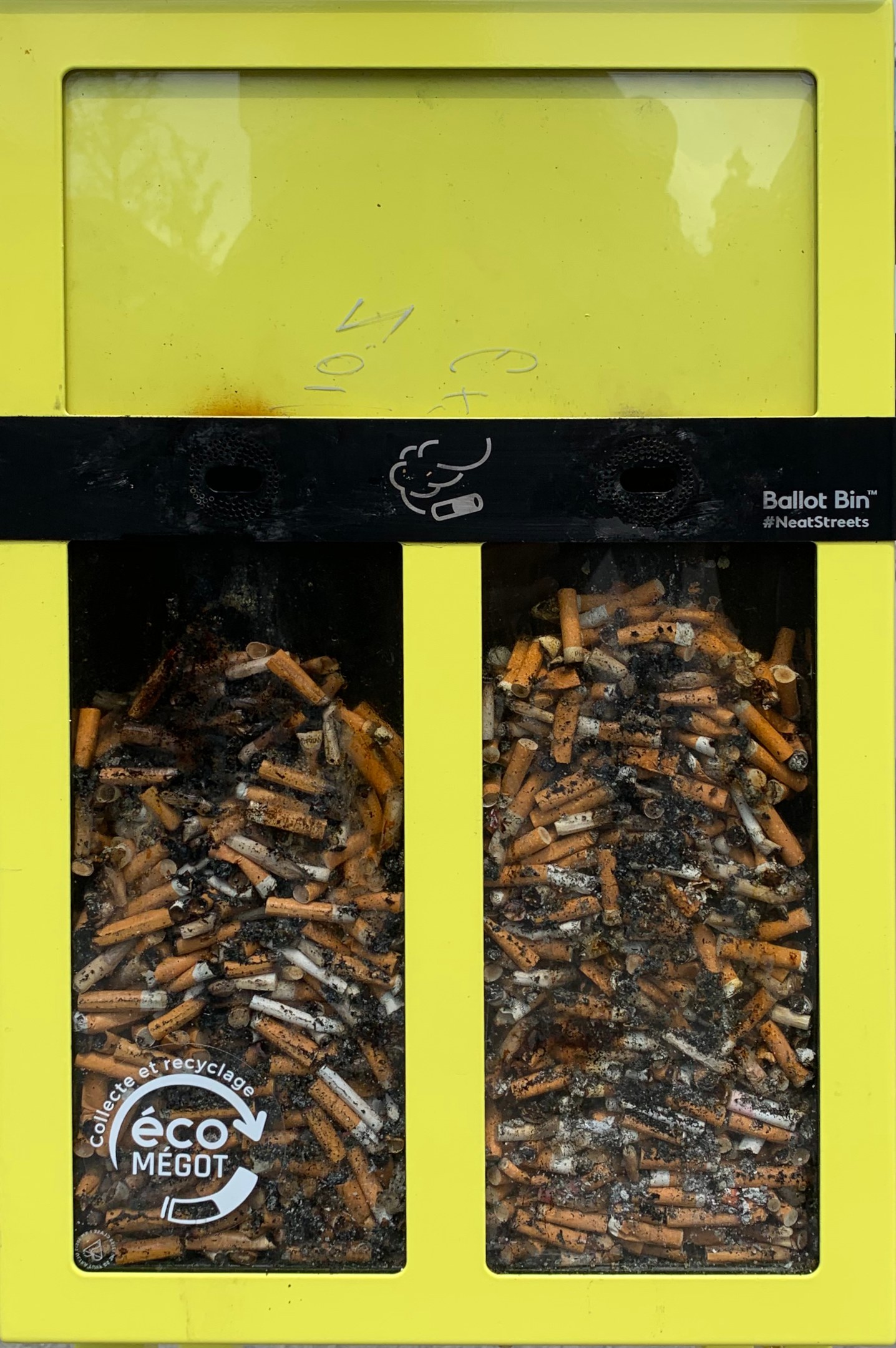 Ein strahlend gelber Recyclingmüllcontainer voller Zigarettenstummeln auf einem Gehweg, beschriftet für die Entsorgung.