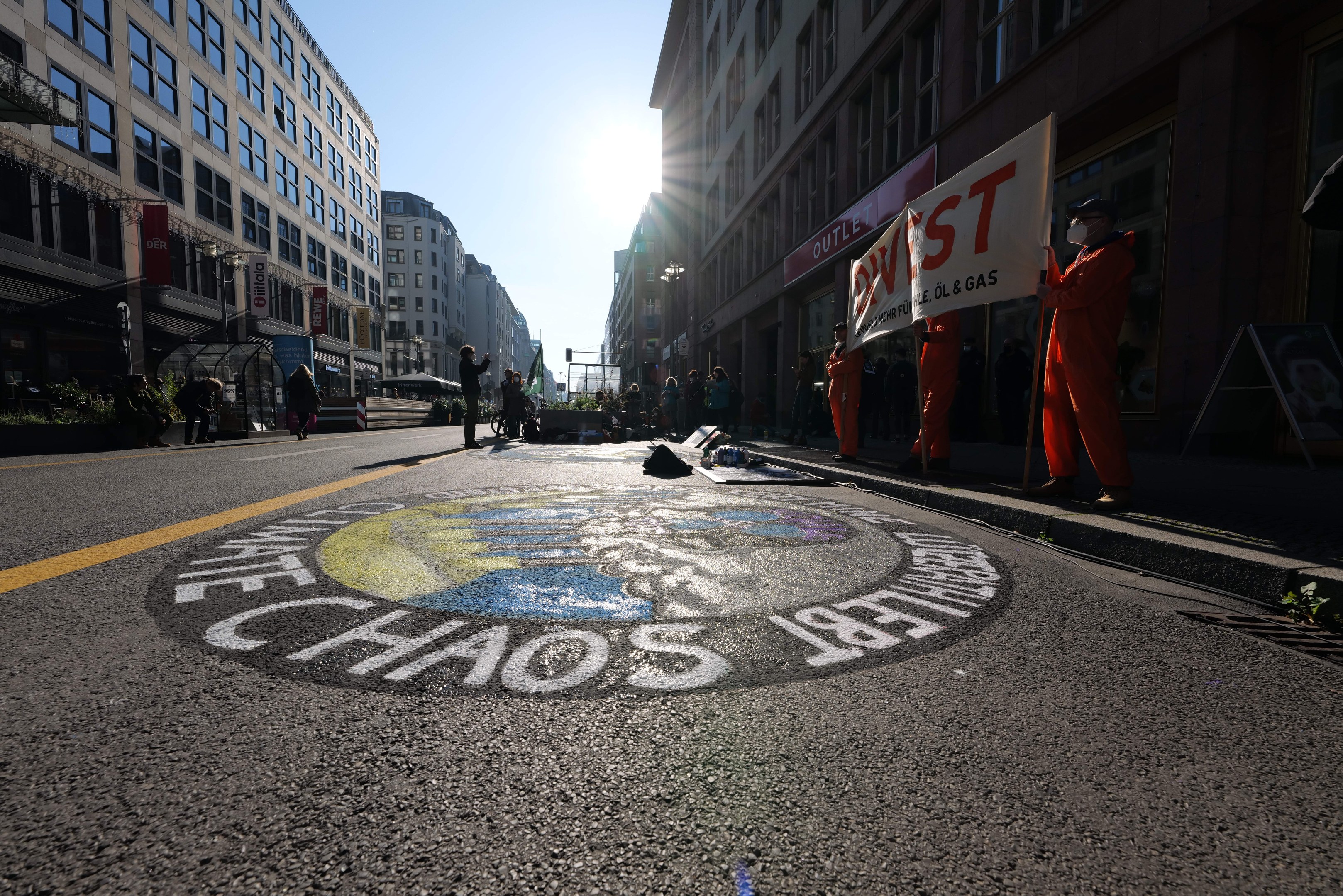 Eine Gruppe von Menschen mit Schildern und Plakaten steht auf einer Straße, die mit Pflanzen und Gebäuden gesäumt ist, mit "Chaos" auf der Straße dahinter gemalt.