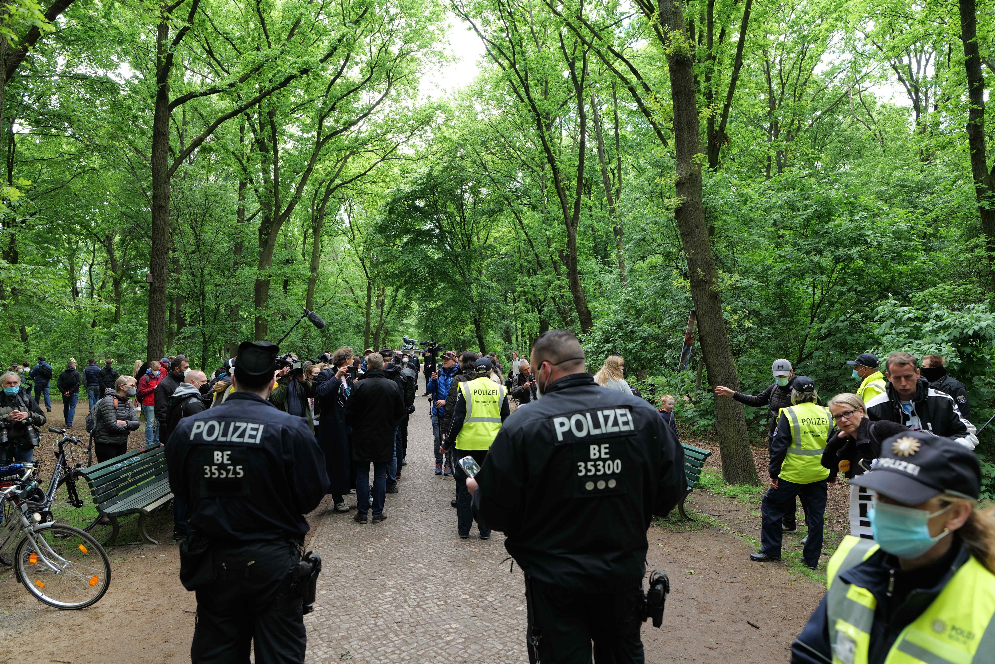 Gruppe von Polizeibeamten vor einer Menge bei einer Anti-Terror-Demonstration in Berlin, mit Fahrrädern und einer Bank im Vordergrund und Bäumen im Hintergrund.