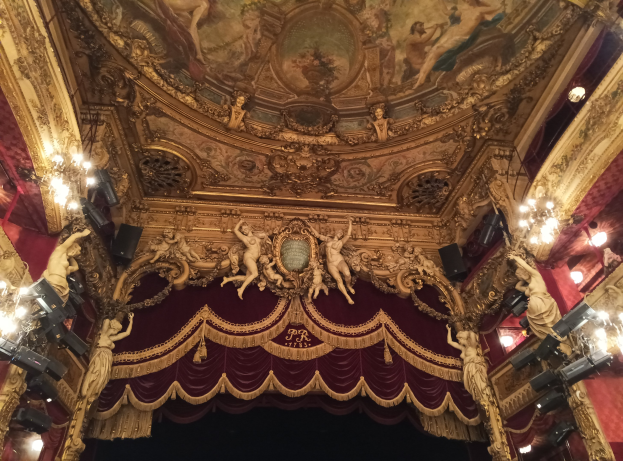 Innere eines Theaters mit rotem Vorhang, Kronleuchtern, Skulpturen, Lichtern und einer Deckenmalerei, die eine Bühne mit ein paar Menschen am Boden zeigt.