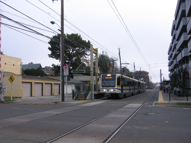 Straßenbahn fährt durch eine Straße mit hohen Gebäuden, Strommasten mit Drähten, Fußgängern auf dem Gehweg, Bäumen und einem klaren blauen Himmel im Hintergrund.