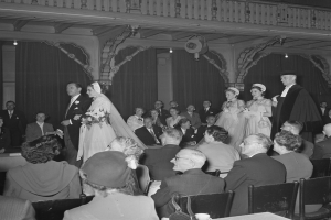 Ein Schwarz-Weiß-Foto einer Hochzeitszeremonie in einem großen Raum mit vielen sitzenden und stehenden Gästen, einige halten Blumensträuße, und Tische mit Tassen und anderen Gegenständen, mit Vorhängen und Deckenleuchten im Hintergrund.