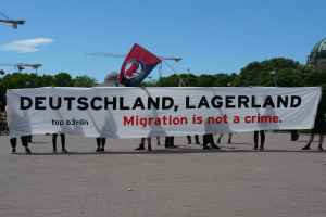Gruppe von Menschen mit einem Banner, auf dem "Deutschland, Lagerland Migration ist kein Verbrechen" steht, und einer Flagge, im Hintergrund sind Laternenpfähle, Straßenlaternen, Bäume, Kräne, ein Gebäude und ein bewölkter Himmel zu sehen.