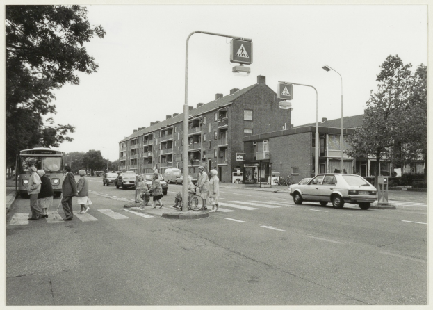 Ein Schwarz-Weiß-Foto von Menschen, die eine Straße überqueren, mit Fahrzeugen, Fußgängern auf dem Gehweg, einem Radfahrer, einem Schild, Straßenlaternen, Bäumen, Gebäuden und einem bewölkten Himmel.