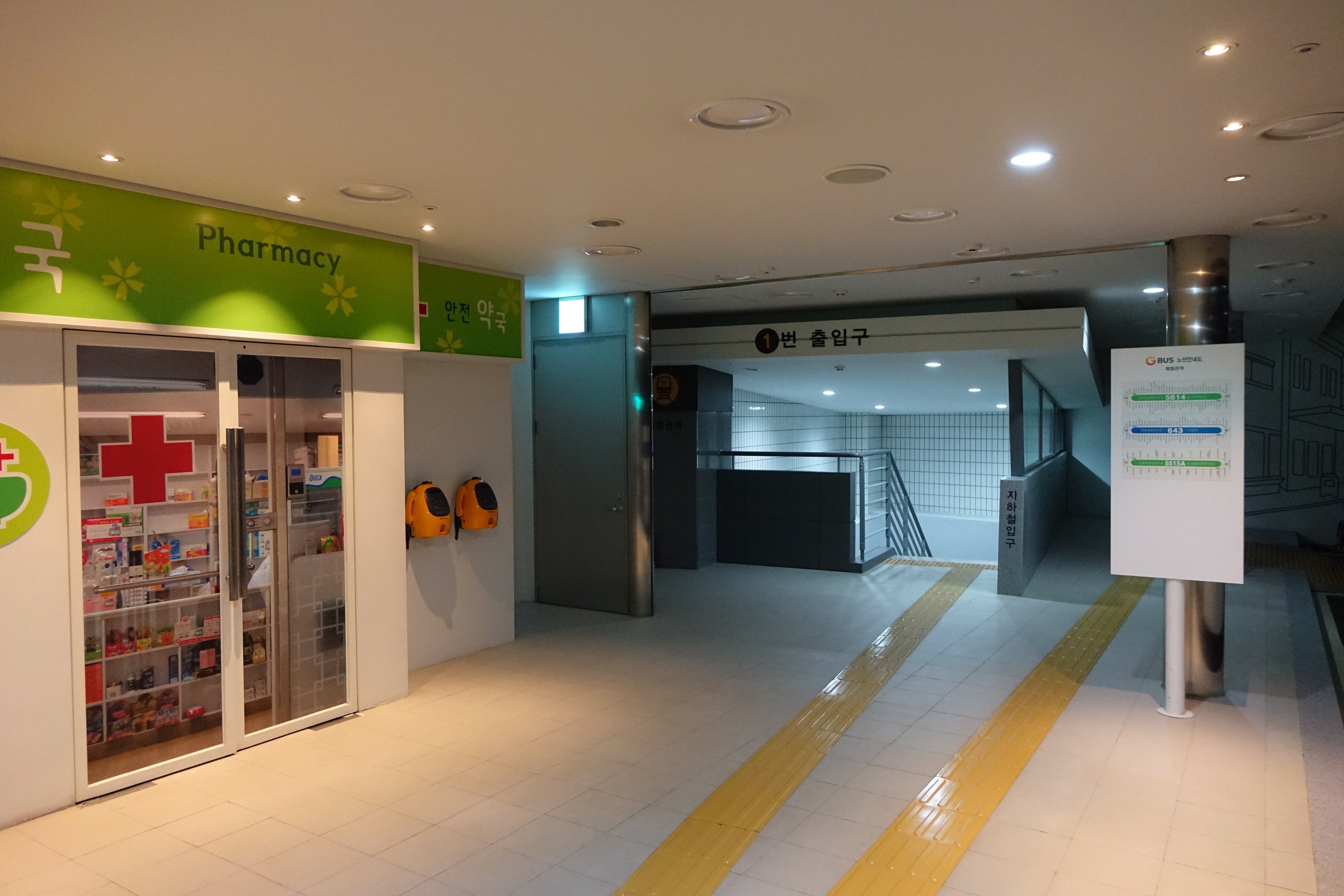 Apothekeneingang mit einem "Apotheke"-Schild auf der linken Seite, einer Tafel mit Text auf der rechten Seite, beleuchteten Lampen darüber und Gegenständen im Hintergrund.