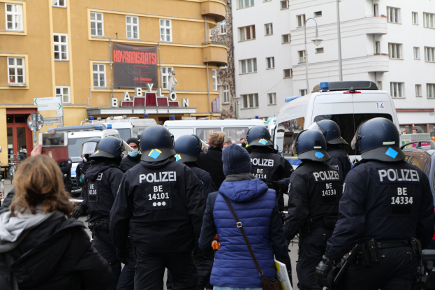 Polizeibeamte in Uniform vor einer Menge von Helmträgern während einer Demonstration in Berlin, mit Fahrzeugen, Gebäuden und einer Kamera im Hintergrund.