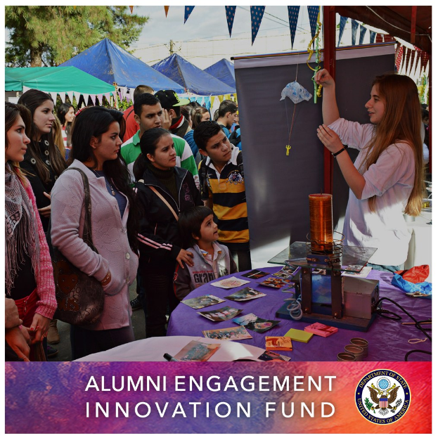 Gruppe von Menschen um einen Tisch versammelt, auf dem ein Schild mit der Aufschrift "Alumni Engagement Innovation Fund" liegt, mit verschiedenen Gegenständen auf dem Tisch, vor einem Hintergrund aus Zelten, Bäumen und einem klaren blauen Himmel, einschließlich Text und Logo unten.