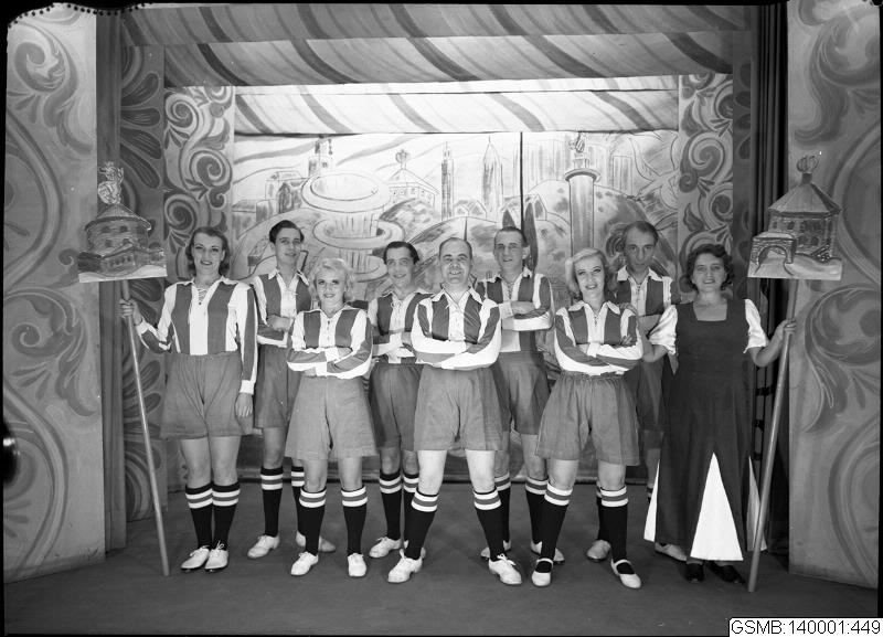 Eine Gruppe lachender Frauen in einem 1940er-Fußballteam posiert auf einer Bühne, jede hält einen Stock in der Hand, mit einer bemalten Wand im Hintergrund.
