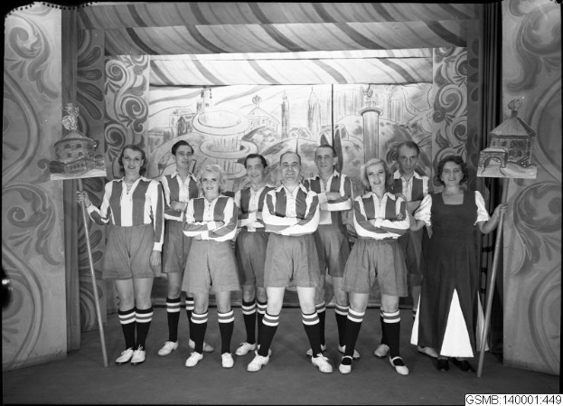Eine Gruppe lachender Frauen in einem 1940er-Fußballteam posiert auf einer Bühne, jede hält einen Stock in der Hand, mit einer bemalten Wand im Hintergrund.