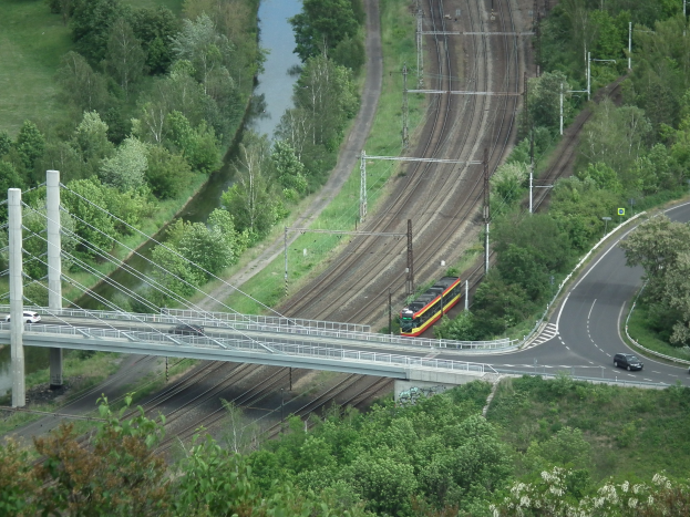 Ein Zug überquert eine Brücke über einen Fluss, mit Fahrzeugen auf der Straße darunter, Strommasten und -leitungen entlang der Brücke und Bäumen und Gras im Hintergrund.