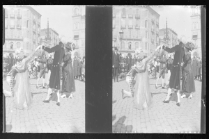 Schwarzes und weißes Foto von Menschen in Kostümen, die auf einer Straße während eines Faschingsumzugs tanzen, umgeben von Gebäuden, Straßenlaternen und Lichtern unter einem klaren Himmel.