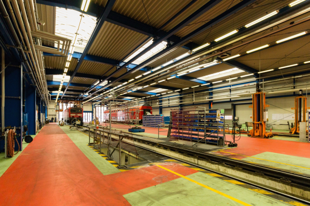 Großer Fabrikinnenraum mit rotem Teppich, Eisenbahnschienen, Geländern, Maschinen, Masten, Lichtern und einem im Hintergrund sichtbaren Zug.