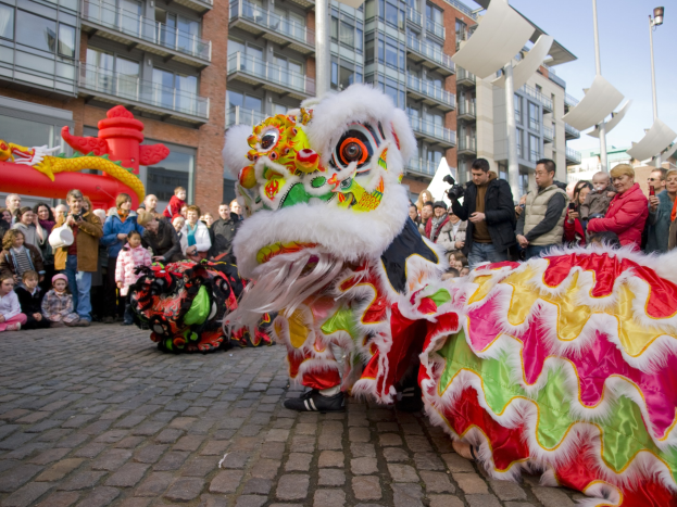 Lebendige Feier des chinesischen Neujahrs in Amsterdam mit Löwen tanzen im Vordergrund und einer Menschenmenge drumherum, einige halten Kameras, vor Gebäuden, Laternenmasten und einem klaren blauen Himmel im Hintergrund.