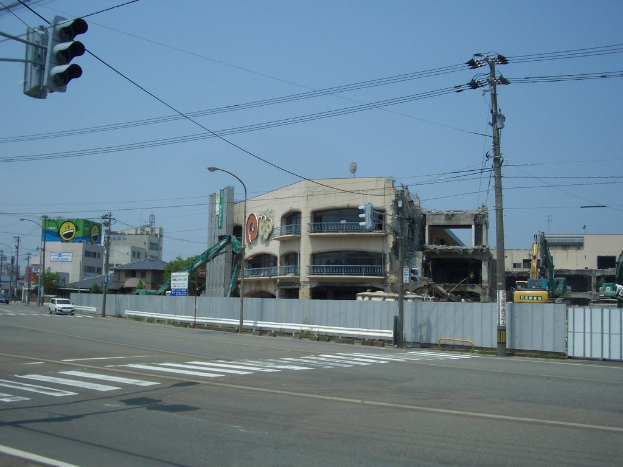 Eine Stadtstraße mit Fahrzeugen, Strommasten, Laternenmasten und einer Ampel auf der linken Seite, Gebäuden, Bäumen und dem Himmel im Hintergrund sowie einem abbruchreifen Gebäude im Vordergrund.