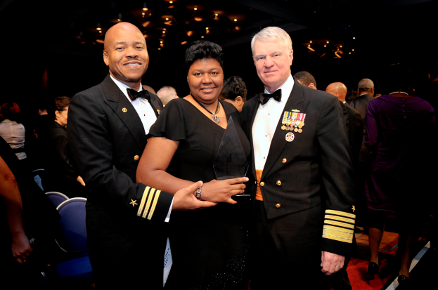 Zwei Männer und eine Frau auf einem roten Teppich; die Frau hält eine Trophäe, alle drei sind formell gekleidet, mit anderen und beleuchteter Decke im Hintergrund.