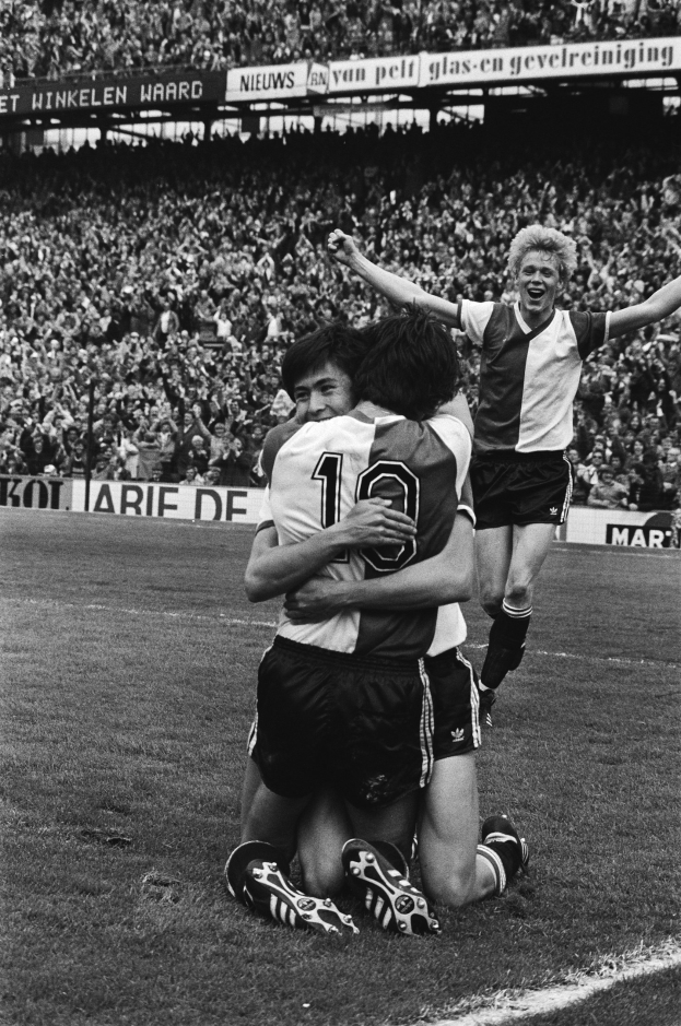 Drei Fußballspieler, die sich nach einem Tor auf dem Boden umarmen, mit jubelnder Menge und schwenkenden Fahnen im Hintergrund, auf einem Schwarz-Weiß-Bild.