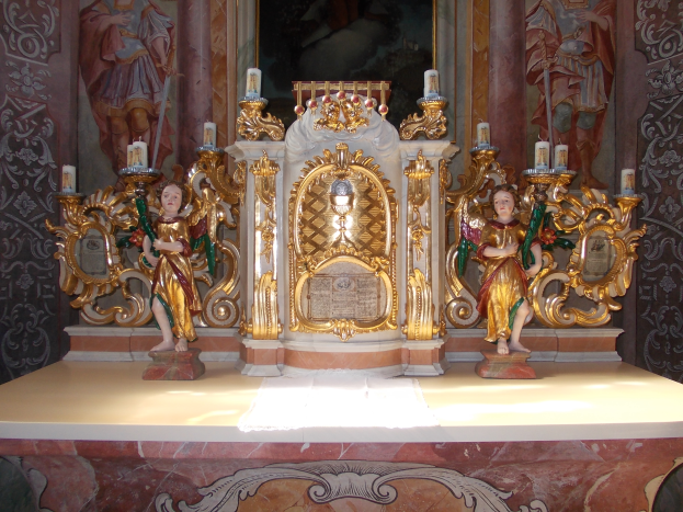 Ein Altar in einer Kirche mit Skulpturen von Menschen und Kerzen auf Ständern, vor einer Wand mit einem Gemälde, die eine feierliche Atmosphäre schaffen.
