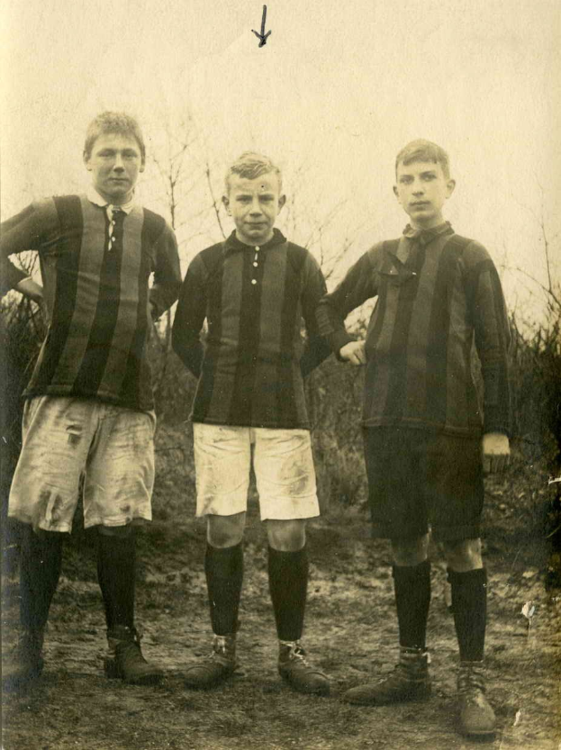 Drei junge Männer in Fußballtrikots auf einem Feld mit Bäumen und einem klaren Himmel im Hintergrund.