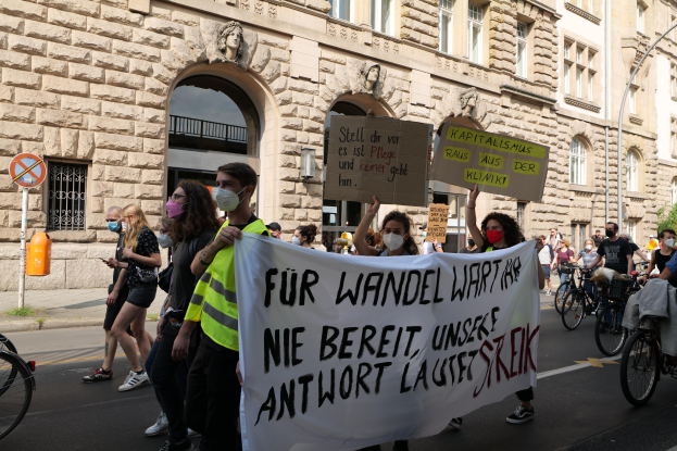 Eine Gruppe von Menschen, die auf einer Demonstration durch die Straße marschieren, einige halten Schilder und andere fahren Fahrráder, im Hintergrund ein Gebäude mit Fenstern, Bogengängen, Pfeilern und Skulpturen, Bäumen und einem Laternenpfahl.