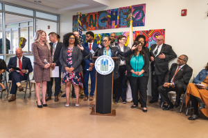 Gruppe von Menschen vor einem Rednerpult mit Mikrofon und einer Tafel mit der Aufschrift "Sancho-Stadtrat tritt dem Stadtrat bei", mit sitzenden und stehenden Teilnehmern, Flaggen, Feuerlöscher und Deckenlampen im Hintergrund.