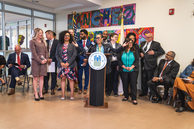 Gruppe von Menschen vor einem Rednerpult mit Mikrofon und einer Tafel mit der Aufschrift "Sancho-Stadtrat tritt dem Stadtrat bei", mit sitzenden und stehenden Teilnehmern, Flaggen, Feuerlöscher und Deckenlampen im Hintergrund.