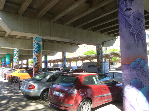 Ein Parkplatz mit zahlreichen geparkten Autos unter einer Brücke, mit Graffiti an den Pfeilern und näheren Gebäuden, mit Bäumen im Hintergrund und einem Van unter den Fahrzeugen.