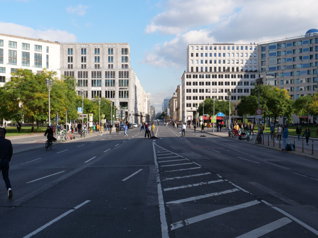 Eine belebte Stadtstraße in Berlin, Deutschland, mit Fußgängern und Radfahrern auf der Straße, umgeben von hohen Gebäuden, Bäumen, Laternenmasten und Schildern unter einem bewölkten Himmel.