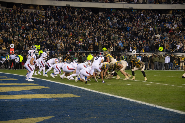 Ein Füssballspiel in einem Stadion mit Spielern in Sportbekleidung und Helmen auf dem Feld, Zuschauern, Geländern und Tafeln mit Text im Hintergrund.