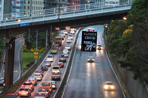 Eine belebte Stadtstraße mit starkem Verkehr, eine Brücke im Hintergrund und ein Banner mit Text in der Mitte der Straße; die rechte Seite zeigt Bäume, Pfosten, Laternen, Schilder und Gebäude.