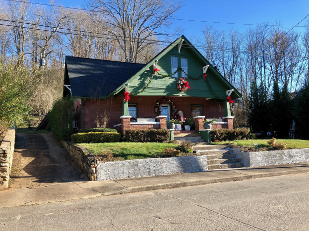 Ein gr├╝nes Haus mit einem Kranz an der Vorderseite, umgeben von Pflanzen, Gras, Treppen, einer Wand, einer Baumgruppe, einigen Dr├Ąhten und einem bew├Âlktem Himmel, gelegen im Herzen der Stadt und zur Miete verf├╝gbar.