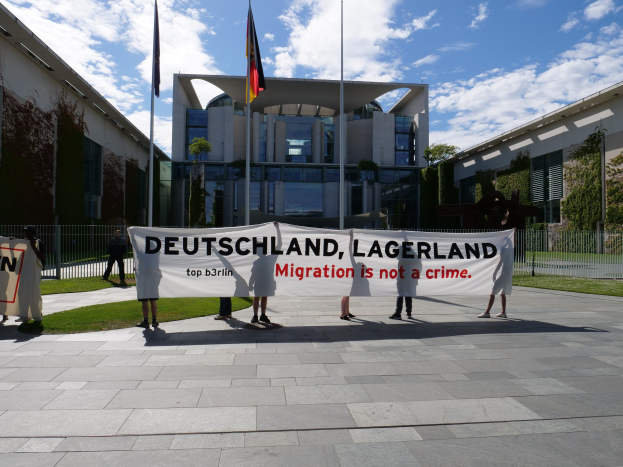 Gruppe von Menschen mit einem Banner "Deutschland, Lagerland Migration ist kein Verbrechen" vor einem Gebäude mit Fenstern, umgeben von Gras, einem Metallzaun, Pflanzen, Bäumen und Fahnenmasten unter einem bewölkten Himmel.