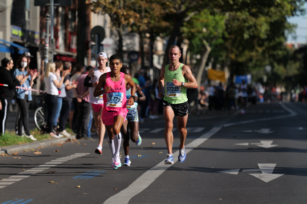 Eine Gruppe von Menschen, die an einem Marathon auf einer Stadtstraße laufen, mit Zuschauern auf der linken Seite, unscharfen Rasen, Bäumen, Gebäuden, Pfosten, Brettern und einem Fahrrad im Hintergrund.
