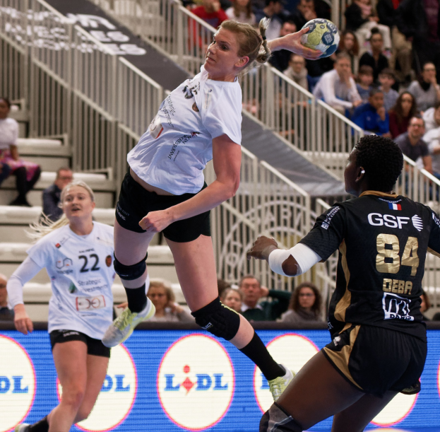 Gruppe von Frauen, die auf einem Platz Handball spielen, mit einem Ball in der Luft, Zuschauern auf Treppen und Geländern und einem "Handball-Weltmeisterschaft 2019 - Tag 2"-Schild im Hintergrund.