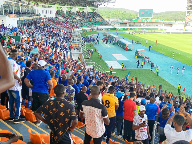 Eine große Menschenmenge steht und sitzt vor einem eingezäunten Fußballfeld mit Hügeln und einem klaren blauen Himmel während der Afrikameisterschaft 2019.