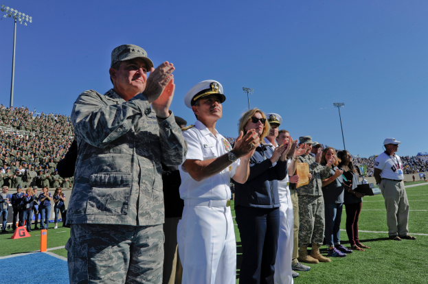 Eine Gruppe von Menschen steht auf einem Fußballfeld und klatscht, mit Zuschauern auf den Tribünen, Flutlichtmasten und einem klaren blauen Himmel im Hintergrund.