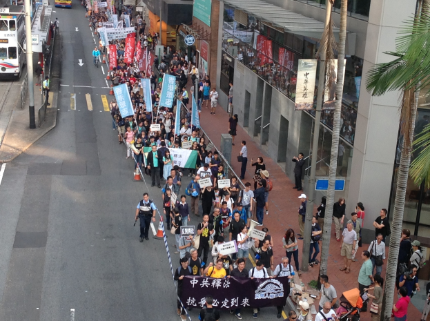 Eine gro├če Gruppe von Menschen marschiert auf einer Stra├če in Hong Kong, mit Protestschildern, mit B├Ąumen, Glasgeb├Ąuden, Fahrzeugen und Straßenschildern im Hintergrund.