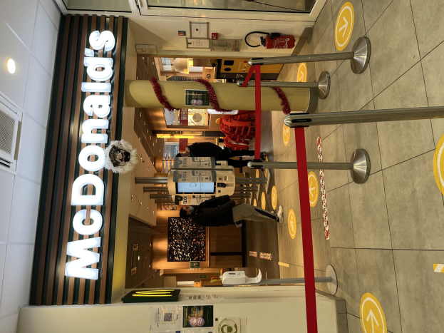 Innenseite eines McDonald's-Restaurants mit einem sichtbaren Schild, Metallstangen mit roten Bändern und einigen Menschen im Vordergrund.