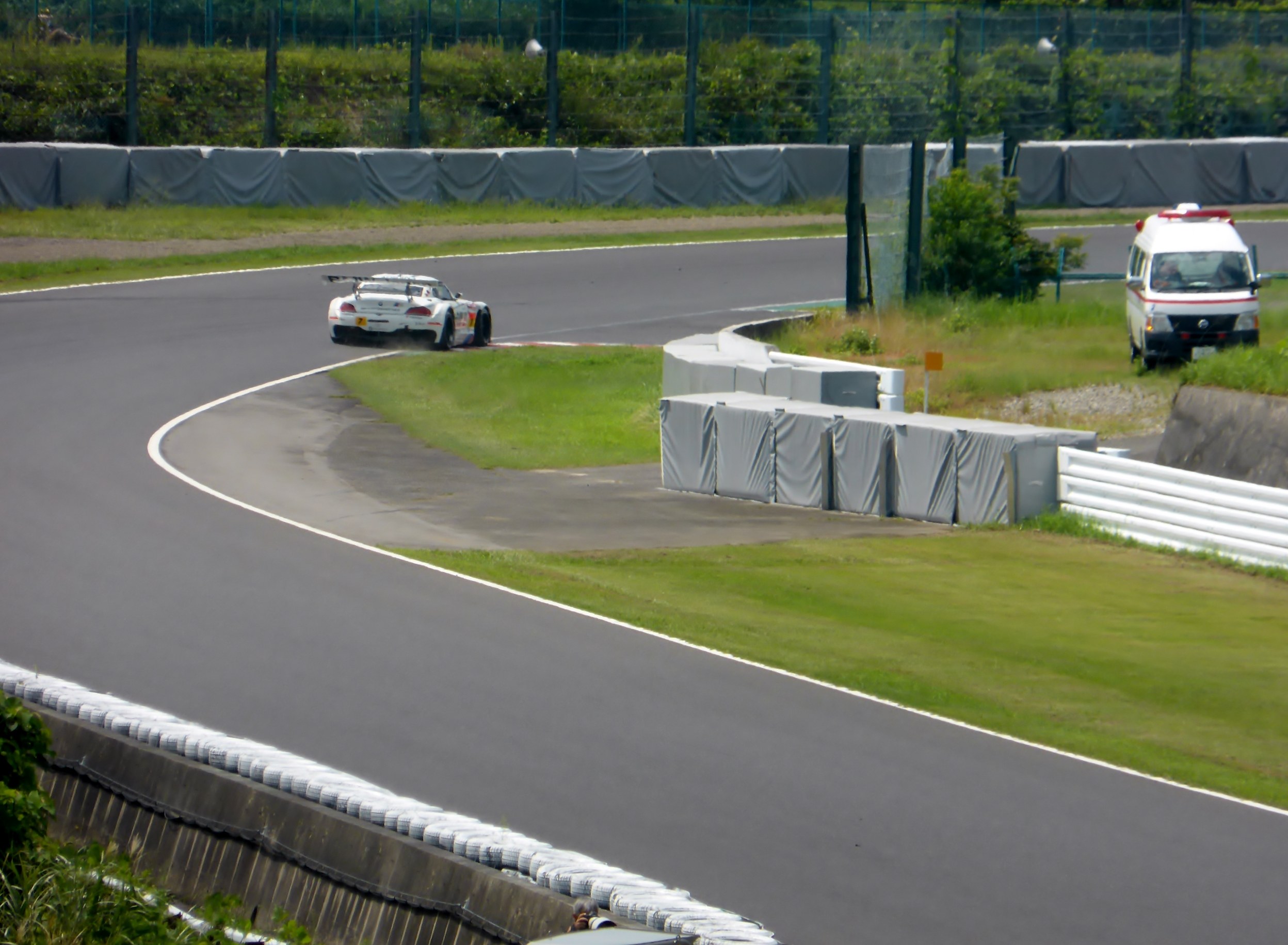 Ein Rennwagen fährt auf einer Rennstrecke mit einem Polizeiwagen im Hintergrund, umgeben von Gras, Pflanzen, Bäumen und einem Zaun, mit einem Fahrzeug auf der rechten Seite und Text unten.