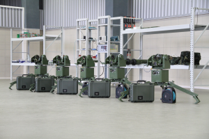 Eine Gruppe von Militärrobotern auf einem Boden mit Koffern und anderen Gegenständen auf umliegenden Gestellen, vor einer Wand mit einem Geländer im Hintergrund.