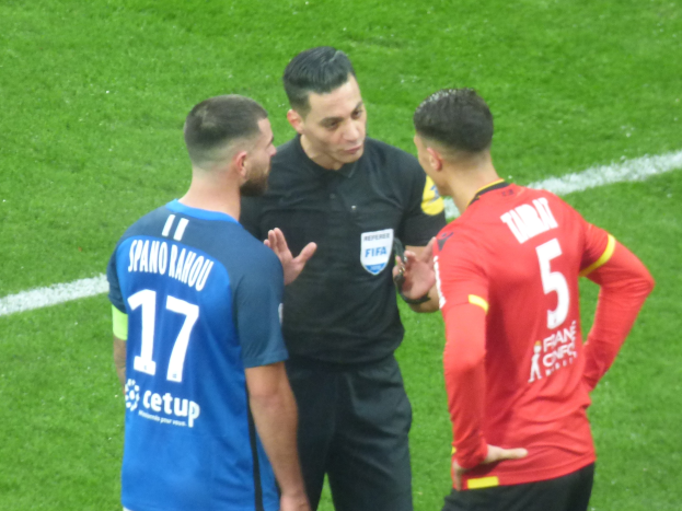 Schiedsrichter spricht mit zwei Männern auf einem Fußballfeld, einer in schwarzer Kleidung und der andere in einem blauen und roten T-Shirt mit Text, beide tragen eine Brille.